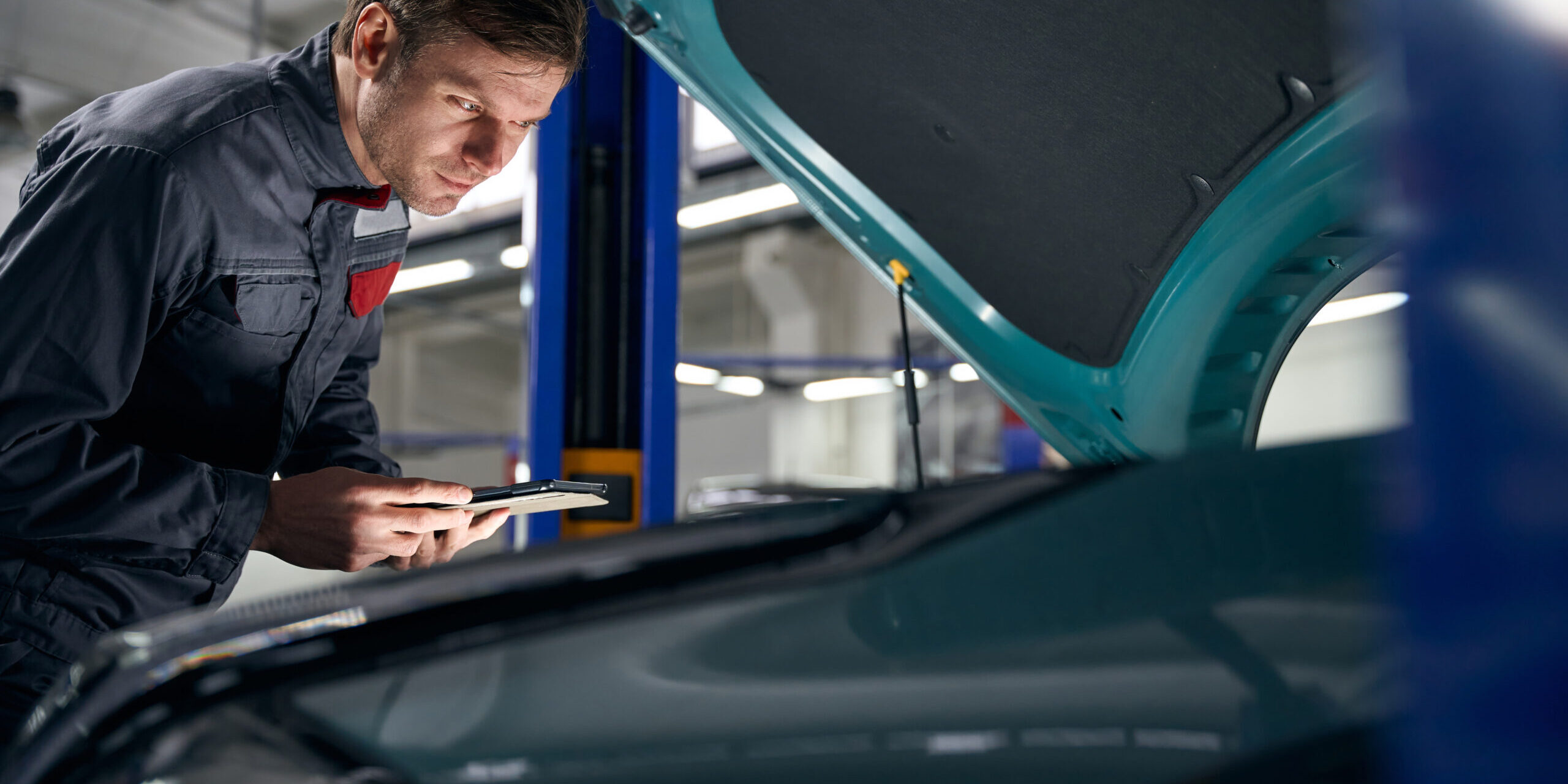 Mechanic checking technical condition of car engine Male in protective clothes standing in tire fitting near car with open hood and holding laptop